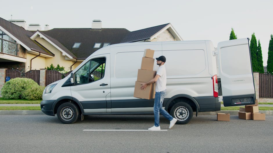 A man wearing a white t-shirt, jeans, and a black cap is loading three cardboard boxes onto the side of a white cargo van parked on a suburban street. The van is positioned parallel to the curb, with its side door open, and additional boxes are placed on the pavement nearby. The background features a row of modern houses with sloped roofs, large windows, and well-maintained gardens, along with a wooden fence and green trees. The scene captures a home relocation or furniture transport process as part of the moving services offered by Man with Van West Ealing, with clear visibility of packed boxes, packaging materials, and moving equipment, emphasizing a professional loading and packing operation.