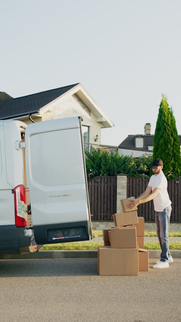 A man in casual clothing loading cardboard boxes into the open rear door of a white moving van parked on a residential street in West Ealing. The boxes are stacked on the pavement beside him, some wrapped in brown packing tape, while he uses both hands to place a box inside the van. The background features a house with a white facade and dark roof, edged by a wooden fence, with well-maintained greenery including a tall, conical shrub. The scene takes place during daylight with clear weather, depicting a home relocation or packing and moving process carried out by Man with Van West Ealing as part of their removals service for the Pitshanger Lane area.