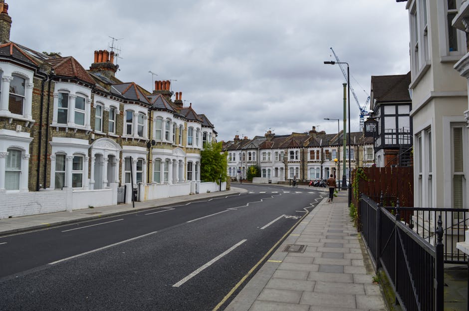 A quiet residential street in West Ealing with row of Victorian-style terraced houses featuring white facades, large bay windows, and red-tiled roofs. The pavement runs alongside the houses with black metal railings in front of some properties. The sky is overcast with grey clouds, and a few pedestrians are visible walking along the sidewalk. On the right side, a white building with multiple windows and a black fence can be seen, while further along the street, a construction crane is visible in the background, indicating ongoing development. The street appears calm and empty of parked vehicles, with a clear, marked road surface. This setting may be relevant to a home relocation or house removals service, where the street provides access for loading or unloading furniture and boxes, supported indirectly by the presence of [COMPANY_NAME] executing a furniture transport or moving logistics operation in the area.