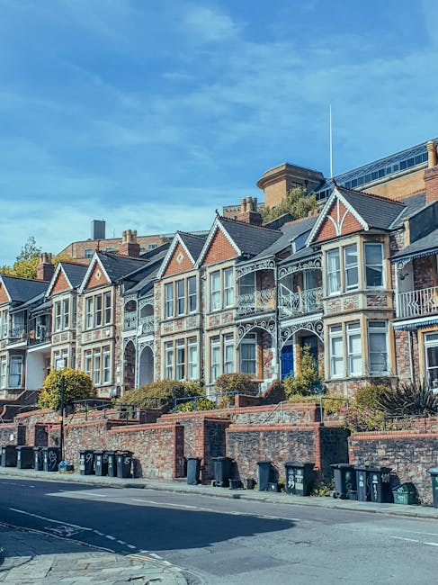 Image depicting a row of Victorian terraced houses with ornate white ironwork balconies and bay windows, situated along a street in West Ealing. The houses are built of brick with some decorative stone details and have pitched roofs with chimneys. The street level features a brick wall with steps leading up to the properties, and black wheelie bins are lined up along the pavement. In the foreground, a section of the road and sidewalk is visible under bright daylight, with some shadows cast from nearby objects, indicating a clear, sunny day. This scene represents a typical residential area involved in house removals and relocation services, where furniture, packed boxes, and moving equipment might be used to facilitate a safe and organized home relocation process. Man with Van West Ealing's house removal services are suited to support such relocations efficiently.