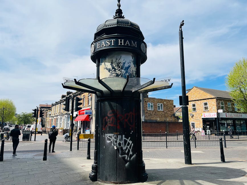 A black, cylindrical underground entrance structure with an ornate, domed top displaying the words 'East Ham' surrounded by decorative elements, situated at a street crossing in a residential area. The structure features a glass panel on the upper part, which appears to have a poster or sign behind it. The surrounding pavement has black metal bollards, and there are pedestrians walking on the sidewalk. Nearby, there is a black lamppost, and behind the underground entrance, there are low brick buildings with retail shops including a nail salon and other stores, and a tree with fresh green leaves indicating spring or early summer. The sky is partly cloudy with large patches of blue, and the overall scene suggests an urban environment, possibly captured during a home relocation or moving process with no immediate furniture or boxes visible.