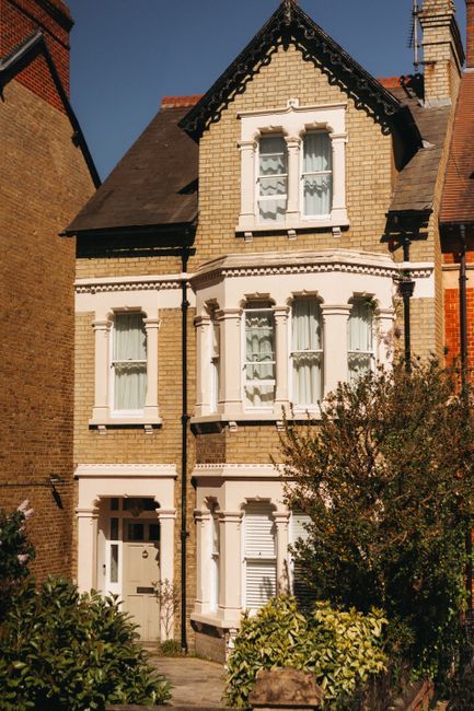 Image depicting a row of Victorian terraced houses with ornate white ironwork balconies and bay windows, situated along a street in West Ealing. The houses are built of brick with some decorative stone details and have pitched roofs with chimneys. The street level features a brick wall with steps leading up to the properties, and black wheelie bins are lined up along the pavement. In the foreground, a section of the road and sidewalk is visible under bright daylight, with some shadows cast from nearby objects, indicating a clear, sunny day. This scene represents a typical residential area involved in house removals and relocation services, where furniture, packed boxes, and moving equipment might be used to facilitate a safe and organized home relocation process. Man with Van West Ealing's house removal services are suited to support such relocations efficiently.